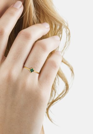 Mano con un delgado anillo de oro que presenta una pequeña piedra verde en forma de trébol, tocando suavemente cabello rubio ondulado contra un fondo blanco.