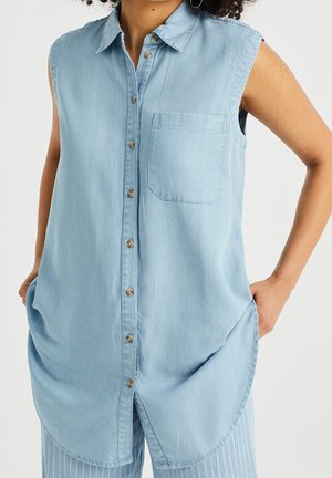 Donna che indossa una camicia di jeans azzurro chiaro senza maniche con bottoni frontali e una tasca sul petto, con le mani posate nelle tasche laterali della camicia.