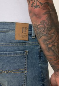 Jeans de mezclilla azul con un parche de cuero que lleva la etiqueta "JP 1880", costuras en tono marrón y un sutil detalle en el bolsillo trasero. Brazo con tatuajes visibles.