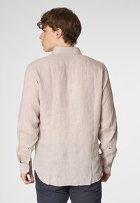 Camicia lunga beige chiaro in tessuto testurizzato, con collo e polsini a bottone, orlo curvo e un leggero lucido.