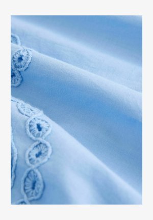 Hellblauem Stoff mit glatter Textur, der an einer Kante über komplexe Stickereien verfügt, die ein zartes, gewelltes Muster erzeugen.