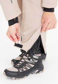 Beige utomhusbyxor med sid dragkedjor, elastiska muddar och kardborredetaljer, parat med svarta och grå vandringsskor med strukturerade sulor.