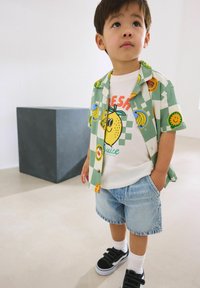 Kind, das ein kurzärmeliges, mintgrünes Shirt mit Obstmustern, ein weißes Grafik-T-Shirt, Jeansshorts und schwarze Sneaker mit Klettverschluss trägt.
