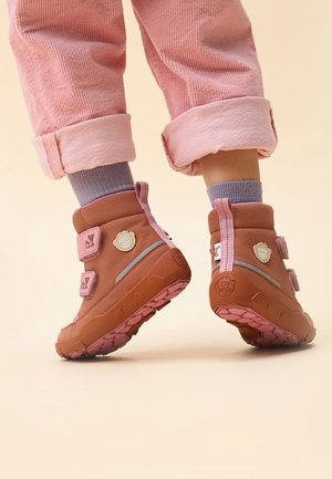Brown insulated boots with pink accents, double Velcro straps, textured soles, and a logo detail on the side, paired with rolled-up pink corduroy pants.