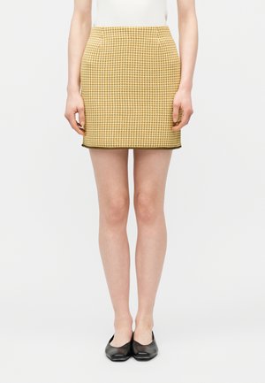 Žena má na sebe mini sukňu v žlto-hnedom vzore psieho zubného kostička, biele tielko bez rukávov a čierne baleríny, stojí na bielom pozadí.