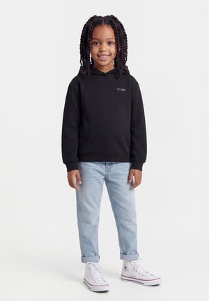 Hymyilevä tyttö, jolla on letitetyt hiukset ja musta huppari, vaaleansiniset ylös käärityt farkut sekä valkoiset korkeavartiset tennarit, seisoo valkoista taustaa vasten.