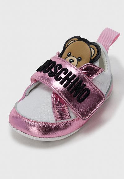 Λευκό δερμάτινο βρεφικό παπούτσι με μεταλλιζέ ροζ λεπτομέρειες, με προσωπάκι αρκούδας στη γλώσσα και ρυθμιζόμενο λουράκι με το «Moschino» μαύρο.