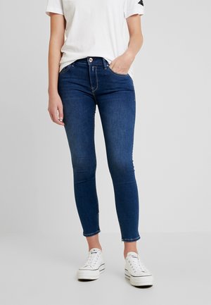 Personne portant un jean skinny bleu foncé, un t-shirt blanc et des baskets à plateforme blanches, debout avec une main dans la poche.