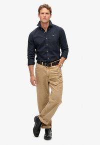 Marineblauw overhemd met een borstzak, beige chino's en zwarte schoenen. Het overhemd heeft een verhoogde kraag en opgerolde mouwen.