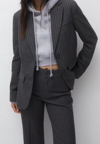 Blazer gris con rayas finas y pantalones a juego, superpuestos sobre una sudadera corta gris. Textura suave y ajuste entallado en todo.