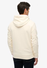 Cremefarvet hættetrøje med blød tekstur, snøre i hætten samt ribstrikkede ærmeafslutninger og kant. Bruges over mørke denimjeans.