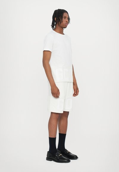 T-shirt bianca in cotone e pantaloni corti coordinati con lettere grandi, abbinati a mocassini neri e calzini blu, su uno sfondo semplice.