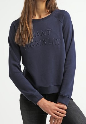 Woman wearing a navy blue sweatshirt with raised text "GANT MCMXLIX," paired with black jeans, against a plain light background.