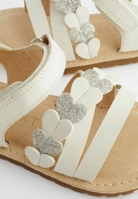 Weiße Sandalen mit einer strukturierten beigen Innensohle, die eine Reihe von glitzernden silbernen Herzen entlang der Riemen aufweisen und eine verstellbare Fesselverschlussschließe haben.