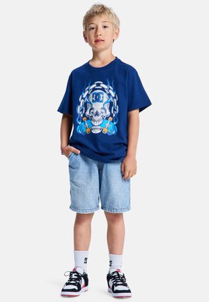T-shirt bleu marine avec un motif de crâne et une chaîne ; associé à un short en jean bleu clair et des baskets noires avec des touches blanches.