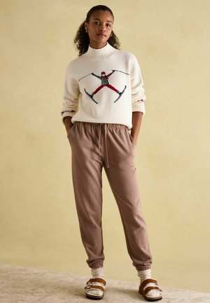 Cream knit sweater with ski graphic, ribbed collar; tan jogger pants with side pockets; brown sandals with white socks.