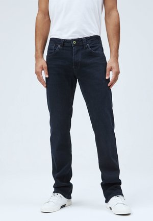 Mężczyzna ubrany w ciemnoniebieskie jeansy, białą koszulkę i białe sportowe buty, stoi z rękami swobodnie opuszczonymi wzdłuż ciała na jednolitym tle.