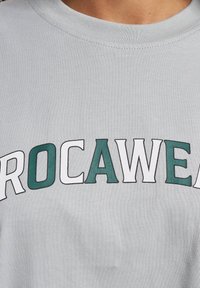 Grå bomull t-skjorte med "PROCAWEAR" i store grønne og hvite bokstaver, rund hals og korte ermer. Glatt tekstur.