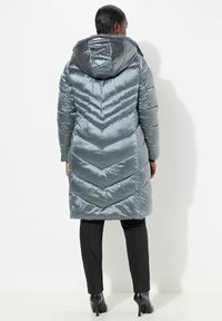 Cappotto lungo imbottito in tessuto grigio lucido con motivo a zigzag, dotato di cappuccio e tasche laterali. Abbinato a pantaloni neri aderenti e tacchi.