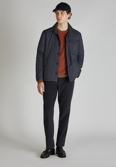 Granatowa lekka kurtka zapinana na guziki, z kieszenią z przodu, zestawiona z swetrem w kolorze rdzy i ciemnymi spodniami. Czarne buty dopełniają stylizację.