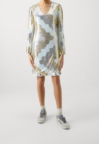 Lesklé šaty s výstřihem do V, zdobené vlnitým vzorem v stříbrné, zlaté a světle modré barvě. Dlouhé rukávy, do délky kolen, doplněné šedými teniskami.
