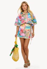 Chemise à motifs floraux colorés et jupe enveloppante assortie, avec des bords festonnés. Les accessoires comprennent un sac tissé jaune et un foulard à motifs.