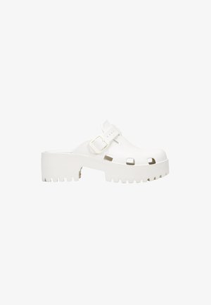 White platform clogs with open toe and cut-out details, featuring a strap with a rectangular buckle. Textured sole with notches.