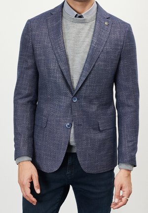 Hombre con blazer azul texturizado sobre suéter gris y camisa azul con cuello, combinado con vaqueros oscuros, de pie con las manos relajadas.