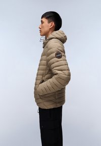 Giacca imbottita beige con cappuccio, caratterizzata da cuciture diagonali e un'etichetta con logo sul braccio sinistro. Abbinata a pantaloni cargo neri.