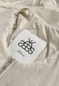 Veste matelassée beige à finition brillante, dotée d'une fermeture éclair argentée et d'une étiquette indiquant « KAREN BY SIMONSEN ». La doublure intérieure est lisse et douce.