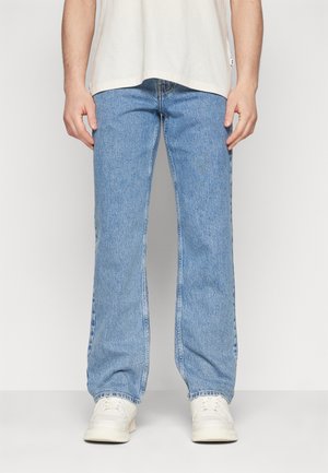 Persoon draagt lichtblauwe rechte jeans, witte sneakers en een beige T-shirt tegen een effen witte achtergrond.