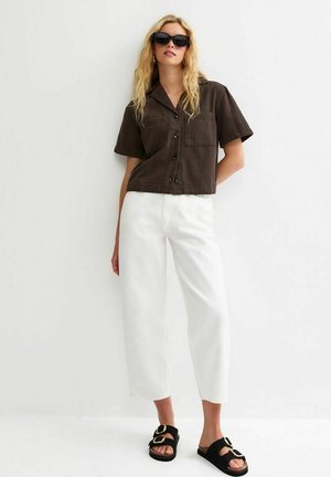 Kobieta o długich blond włosach, nosząca czarne okulary przeciwsłoneczne, brązową koszulę z krótkim rękawem zapinaną na guziki, białe szerokie spodnie oraz czarne sandały z klamrami.