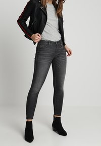 Chaqueta de cuero negra con franjas rojas, camiseta gris, jeans skinny oscuros y botas negras hasta el tobillo. Enfóquese en el estilo ajustado y las texturas.