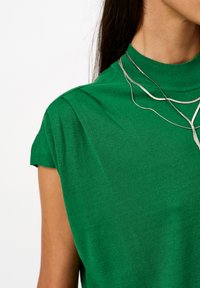 Groene gebreide t-shirt met een hoge halslijn en korte mouwen. Heeft een gestructureerde stof en gelaagde zilveren kettingen.