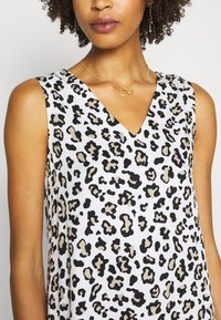 Bílé šaty bez rukávů s výstřihem do V a černobéžovým leopardím potiskem, s jemnou texturou a decentním leskem. Akcent zlatého kruhového náhrdelníku.