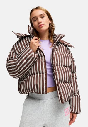 Jeune femme portant une doudoune rayée marron et blanche par-dessus un crop top violet clair et un pantalon de jogging gris avec un texte rose.