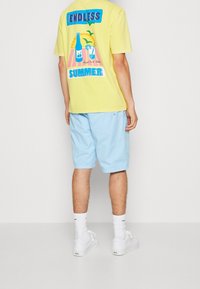 Una camiseta amarilla presenta un diseño gráfico con bebidas y el texto "VERANO INFINITO"; combinada con pantalones cortos azul claro y zapatillas blancas.