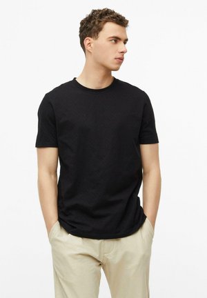 Junger Mann mit lockigem Haar, der ein schwarzes Kurzarm-T-Shirt und beige Hosen trägt, steht mit den Händen in den Taschen und schaut nach rechts.