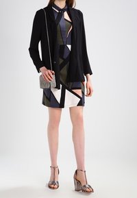 Kvinna klädd i en klänning med geometriskt mönster, svart kavaj, silverfärgade T-sandaler och håller en liten glittrig axelväska.