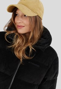 Musta corduroy-puffer-takki, jossa on suuri huppu ja vetoketjusulku. Yhdistetty tekstuuriseen beigeen baseball-lippikseen. Mallilla on laineikkaat hiukset.