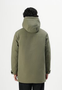 Person mit dunklem, lockigem Haar, der eine olivgrüne, kapuzenartige Winterjacke trägt, mit dem Rücken zu einem schlichten, hellen Hintergrund.