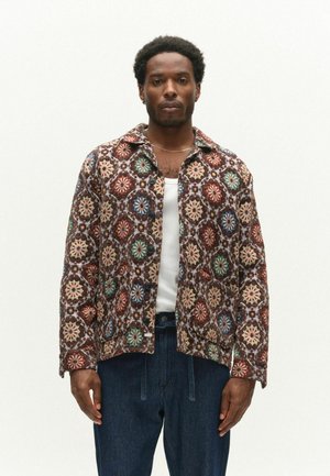 Mann steht und trägt eine geknöpfte Jacke mit mehrfarbigen kreisförmigen Mustern über einem weißen Hemd und dunkelblauer Hose vor weißem Hintergrund.