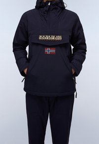 Chaqueta de sudadera con capucha en color marino, con un bolsillo frontal tipo pouch, que presenta un parche con logo y un emblema de la bandera de Noruega. Tiene un corte relajado y bolsillos laterales con cremallera.