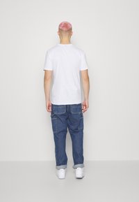 Weißes T-Shirt mit kurzen Ärmeln, lockere blaue Denim-Cargohosen mit Seitentaschen und weiße Turnschuhe, von hinten betrachtet.