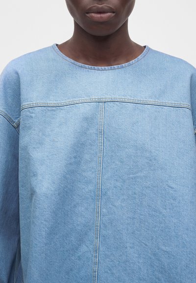 Gros plan d'une personne portant un haut en denim bleu clair avec des détails visibles de coutures sur la poitrine et au centre.