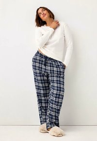 Haut blanc à manches longues associé à un pantalon de pyjama à carreaux bleu marine avec des lignes blanches, une taille élastique, et des chaussons doux beiges.