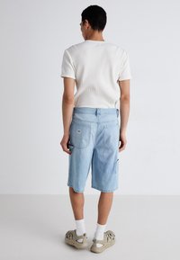Jasnoniebieskie dżinsowe szorty o luźnym kroju, z wieloma kieszeniami i subtelną metką marki, w połączeniu z fakturowanym białym t-shirtem i beżowymi butami.