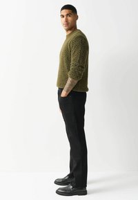 Olivovo zelený chlpatý sveter s čiernymi škvrnami, skombinovaný s čiernymi rovnými nohavicami a čiernymi mokasínami, stojaci pred bielym pozadím.