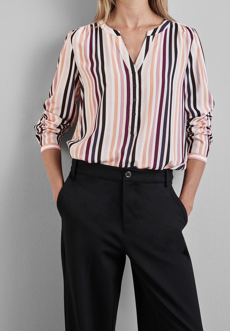 Femme portant un chemisier à manches longues à rayures verticales pêche, rose, noir et blanc avec un pantalon noir ajusté et les mains dans les poches.