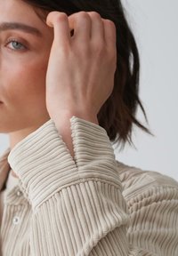 Beige textured top with ribbed detailing, featuring a split cuff design. The fabric appears soft and slightly velvety.